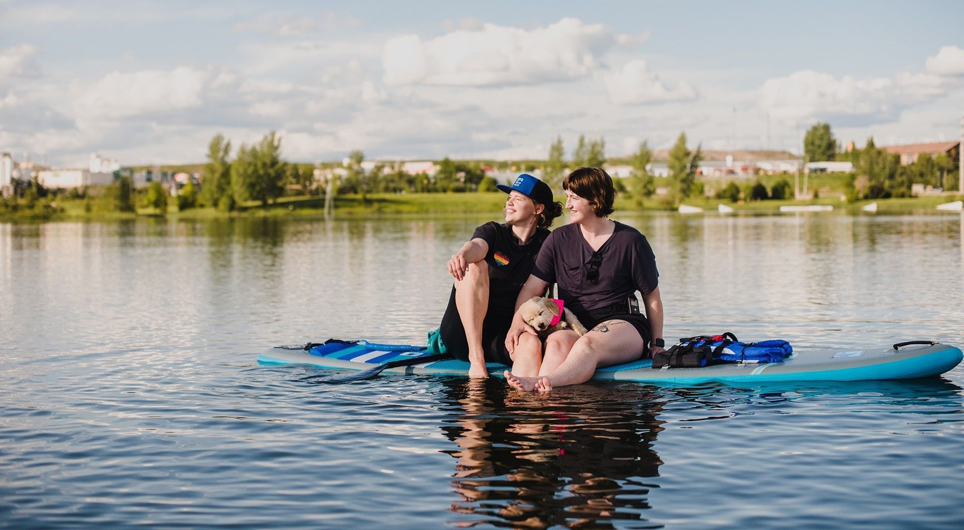 Deux femmes sont assises sur une planche à pagaie, sur un lac. Elles regardent au loin, tout sourire.