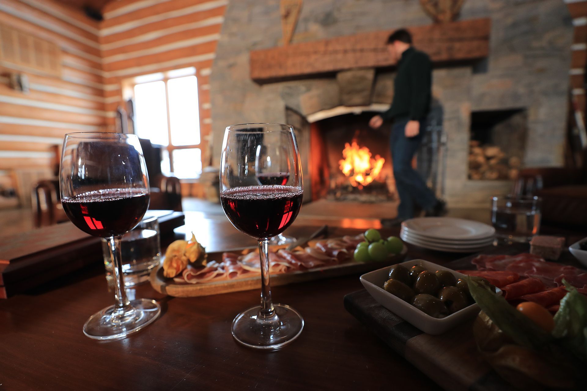 On est dans un chalet. Deux verres de vin et des bouchées sont sur la table. À l'arrière plan, un feu de foyer.