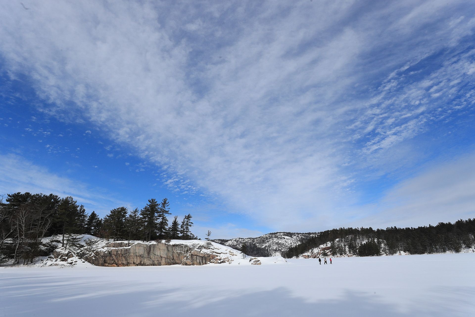 La photo est prise sur un plan d'eau gelé et enneigé. On voit un grand ciel bleu, traversé par un nuage. Des gens se promènent sur la glace.