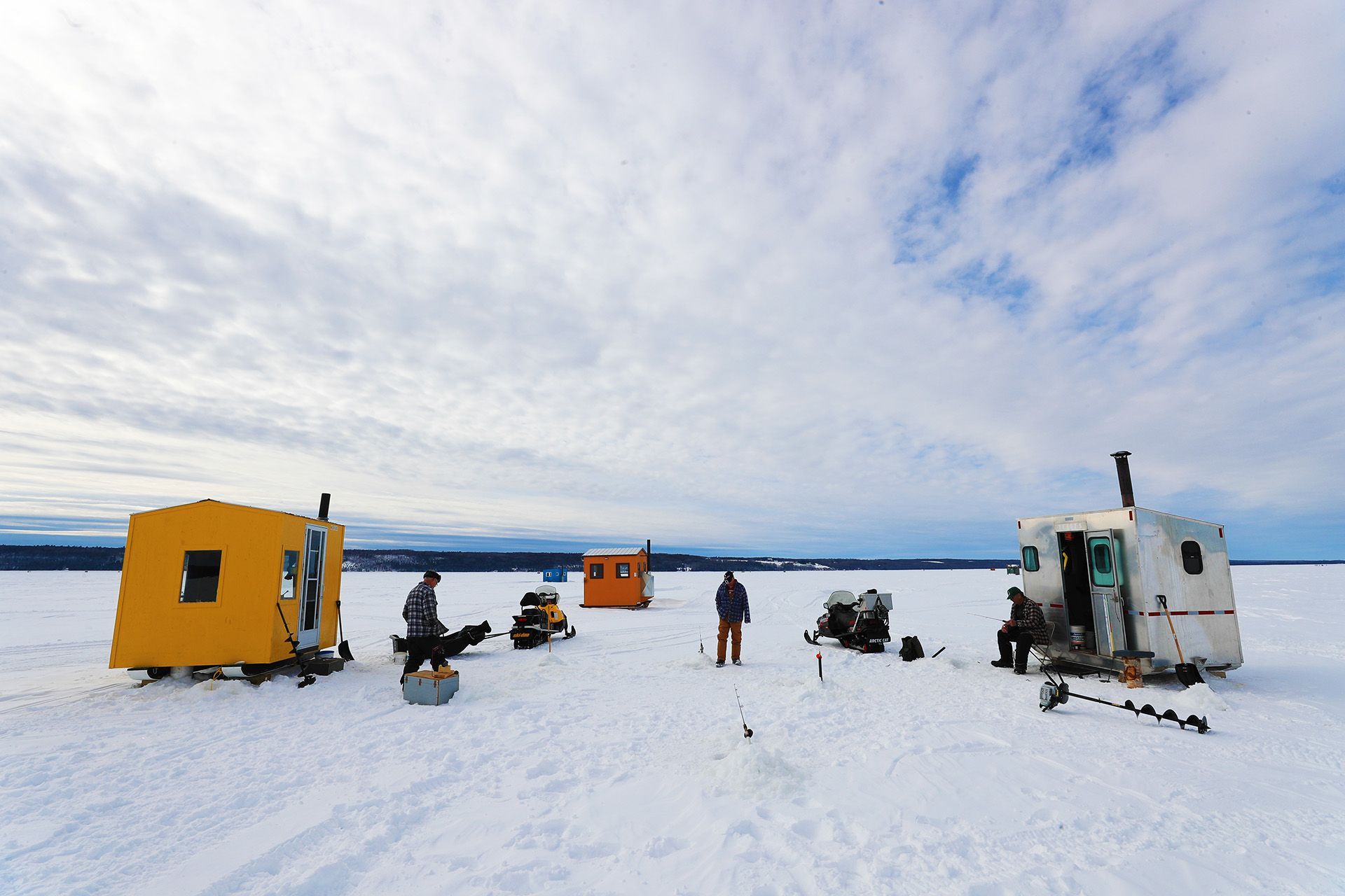 Un village de pêche sur glace, avec trois cabanes et un groupe de quelques pêcheurs, sur un lac. 