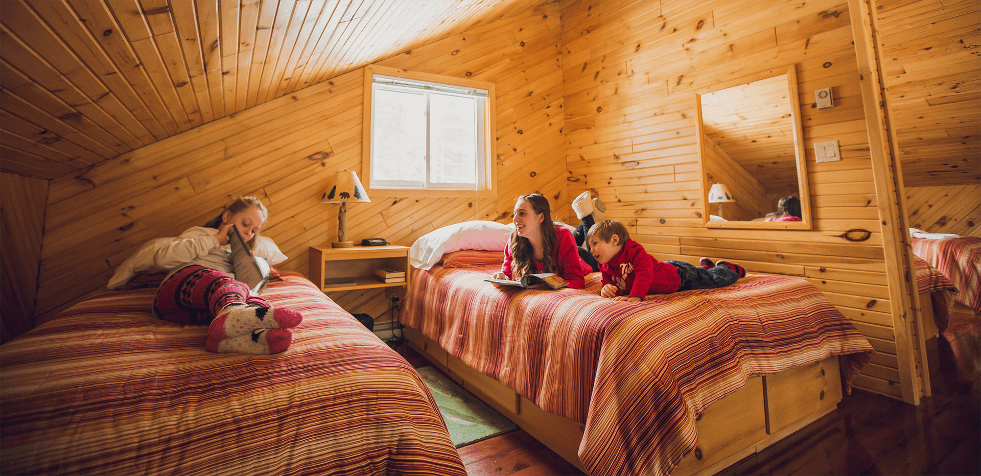 Trois enfants lisent. Ils sont installés sur deux lits dans une chambre lumineuse, aux murs de bois