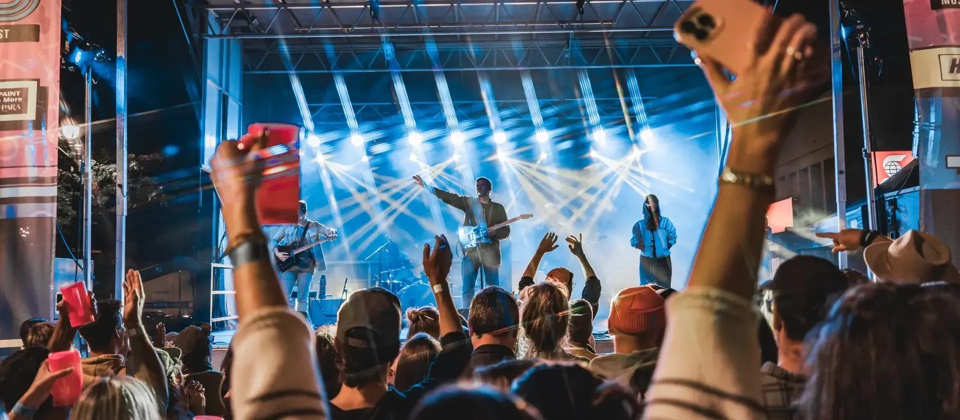 Un festival. La photo est prise depuis la foule. Les gens devant ont les bras en l'air. Les spots illuminent la scène, où se trouvent des musiciens. 