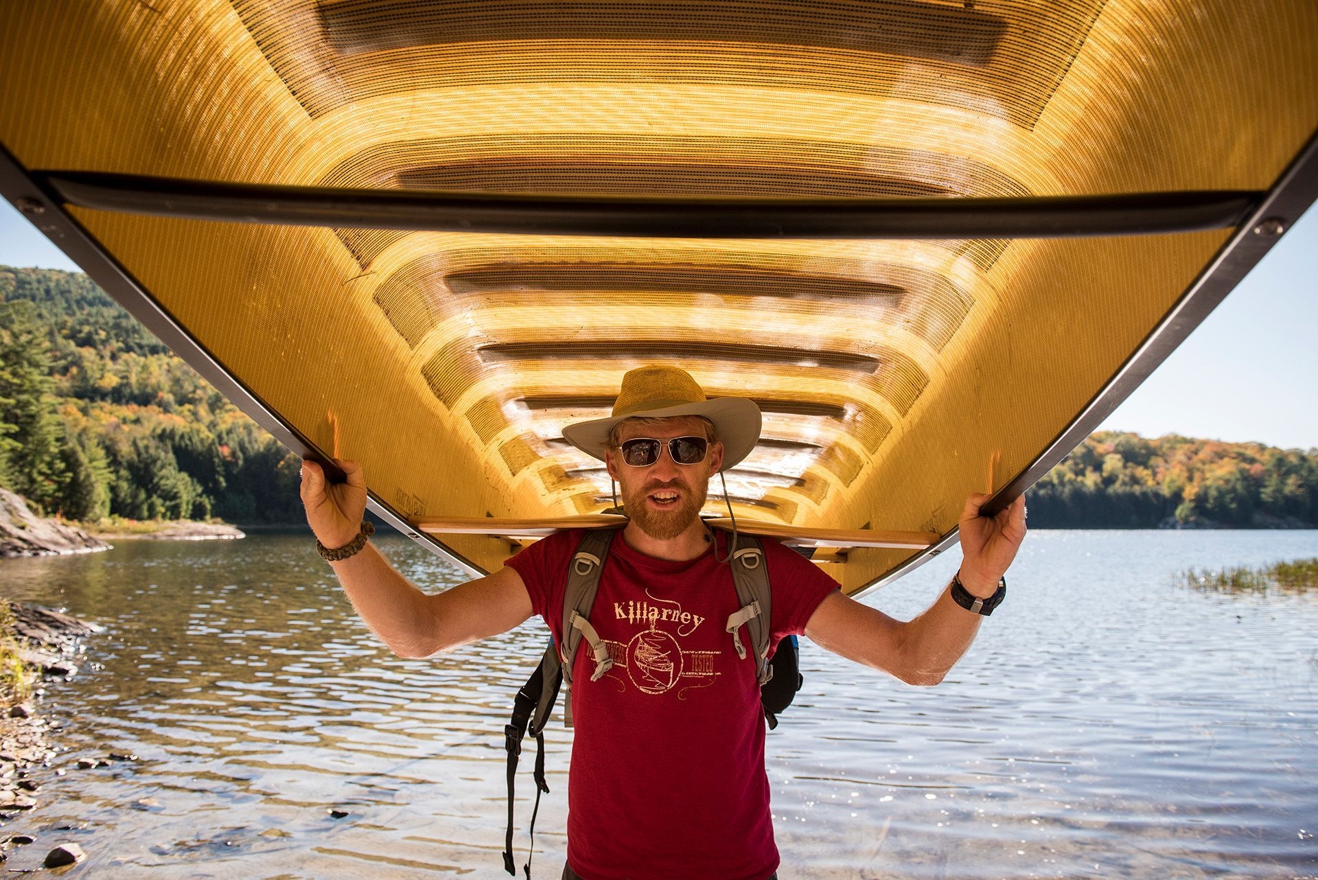 A man in sunglasses and a hat carries a canoe over his shoulders.