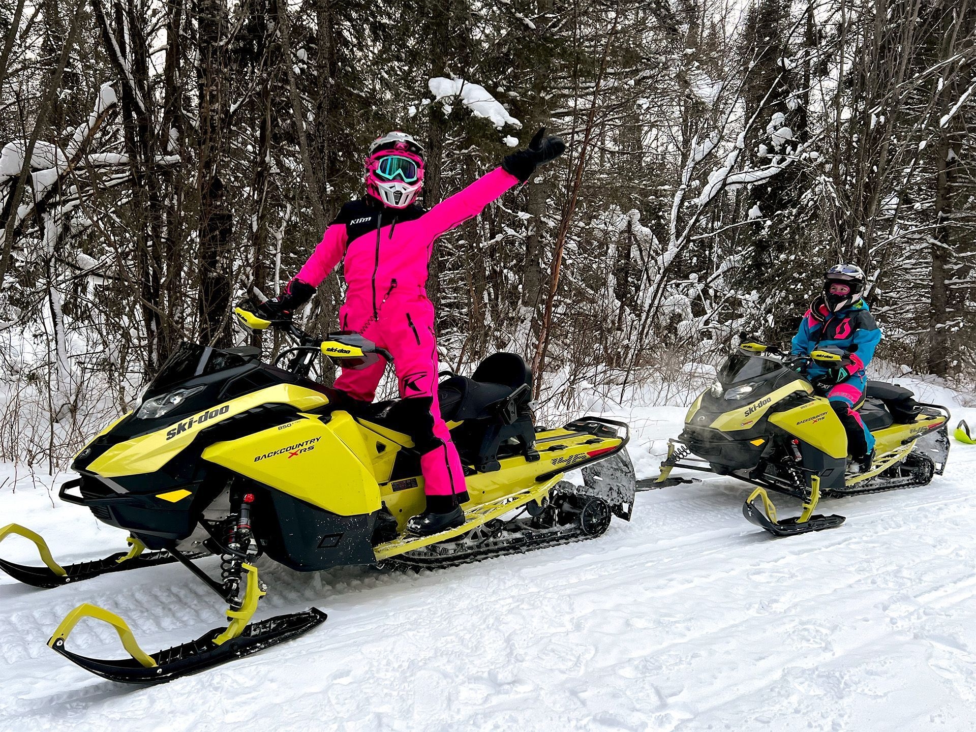 Deux jeunes filles vêtues de combinaisons de neige roses et bleues conduisent des motoneiges jaunes sur un sentier enneigé.