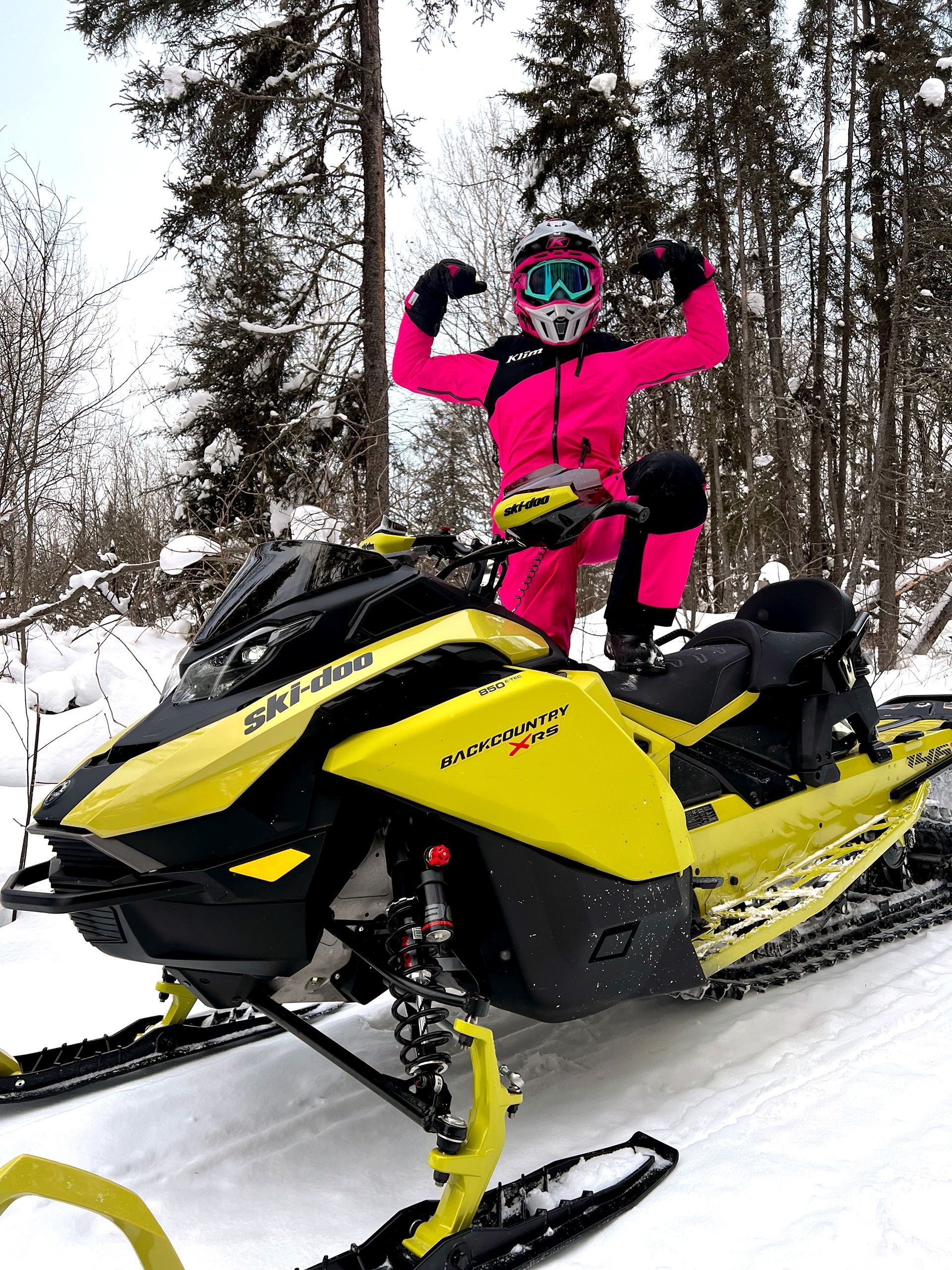 Deux jeunes filles vêtues de combinaisons de neige roses et bleues conduisent des motoneiges jaunes sur un sentier enneigé.