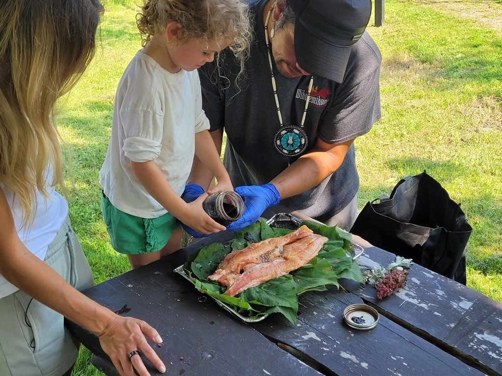 Un jeune enfant aidé d'un chef préparent un filet de saumon, sur des branches de cèdre.