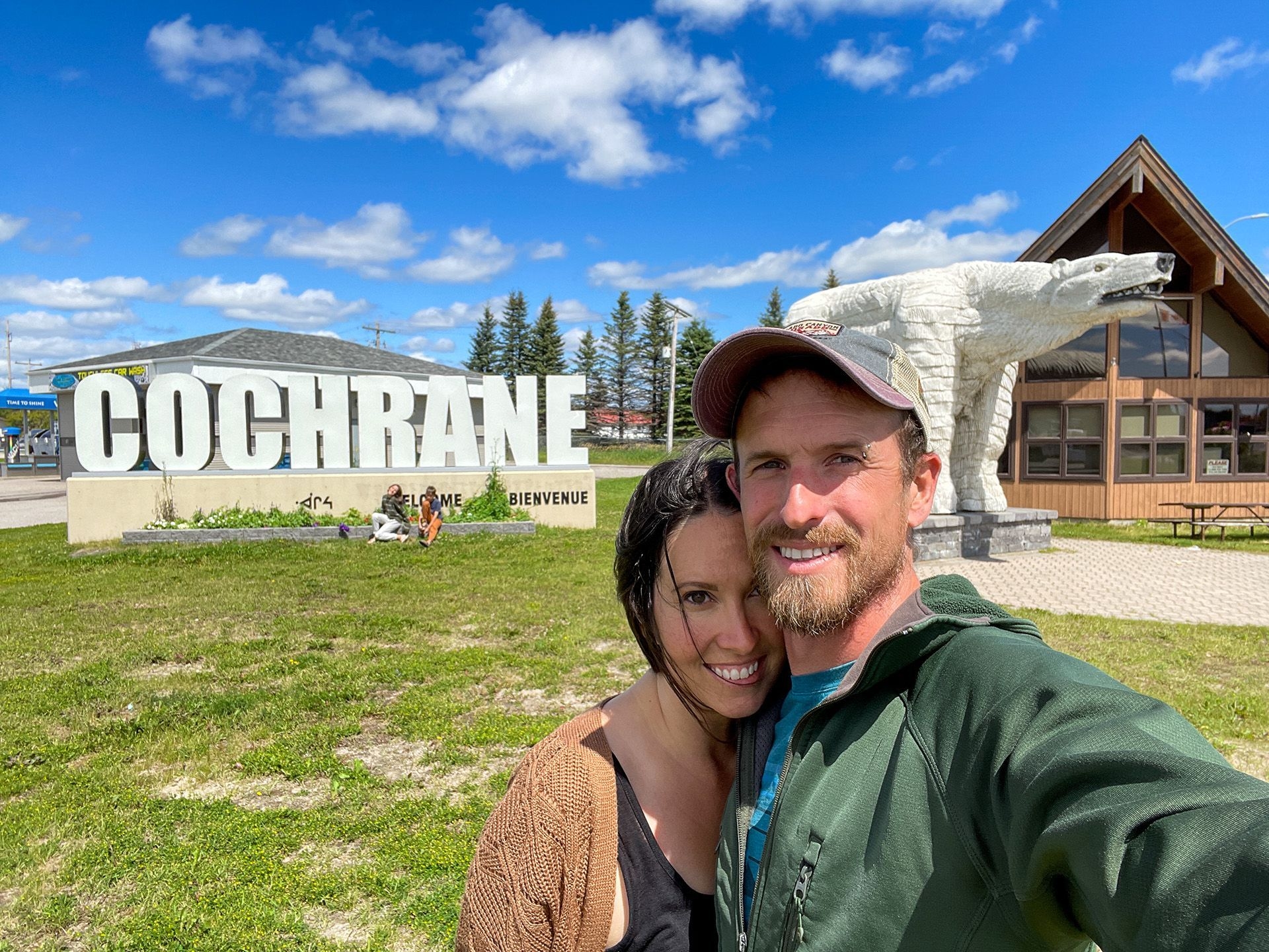 Un couple souriant prend un selfie devant la statue d'ours polaire et le lettrage à l'entrée de Cochrane