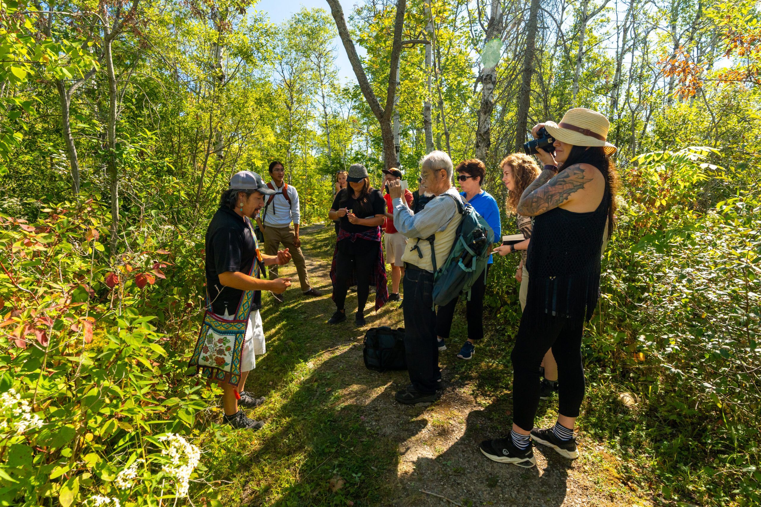 Un groupe écoute attentivement une guide, dans une sentier pédestre, dans une forêt de feuillus. 