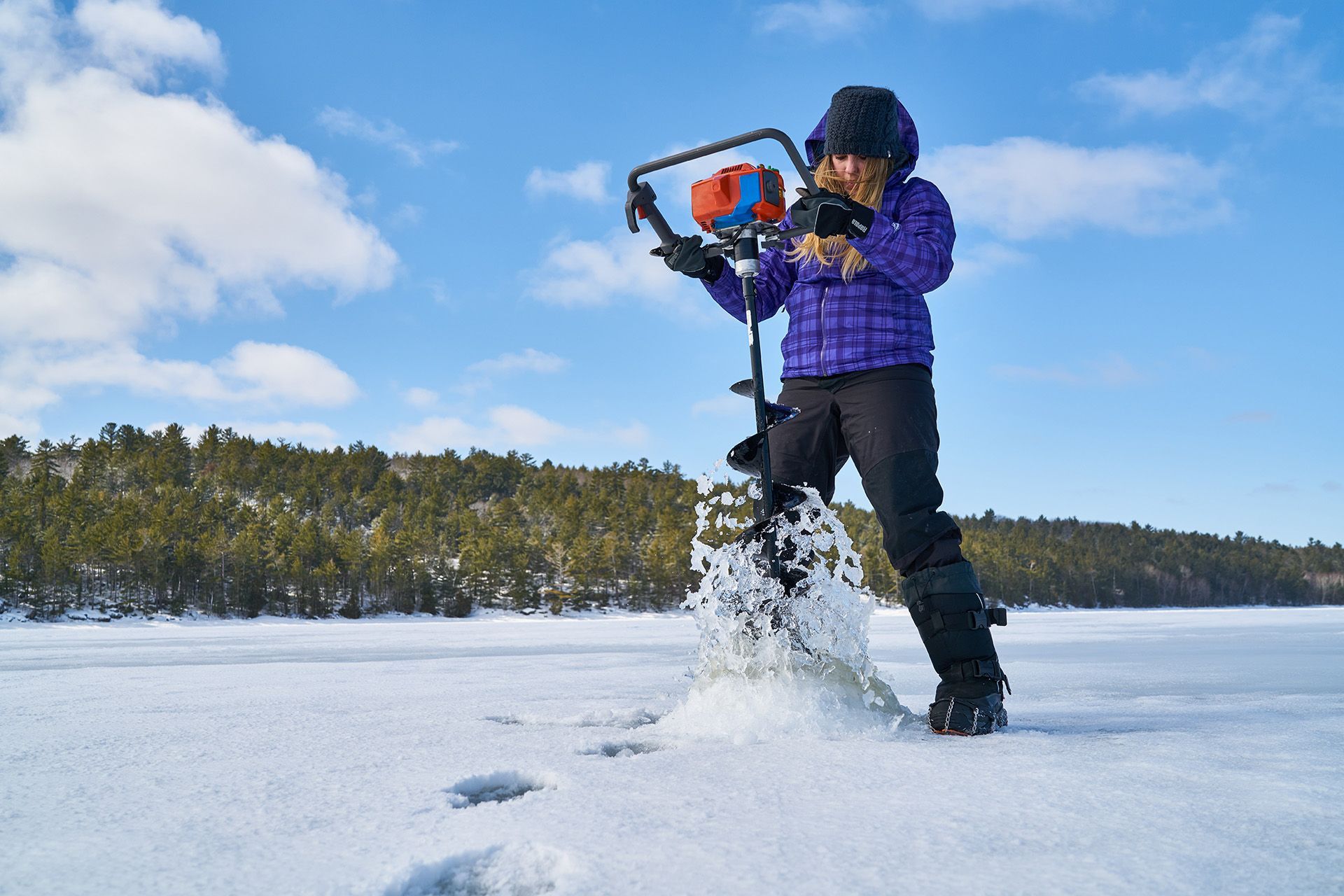 Une personne fait un trou dans la glace, sur un lac. L'eau gicle, aux pieds de la personne. 