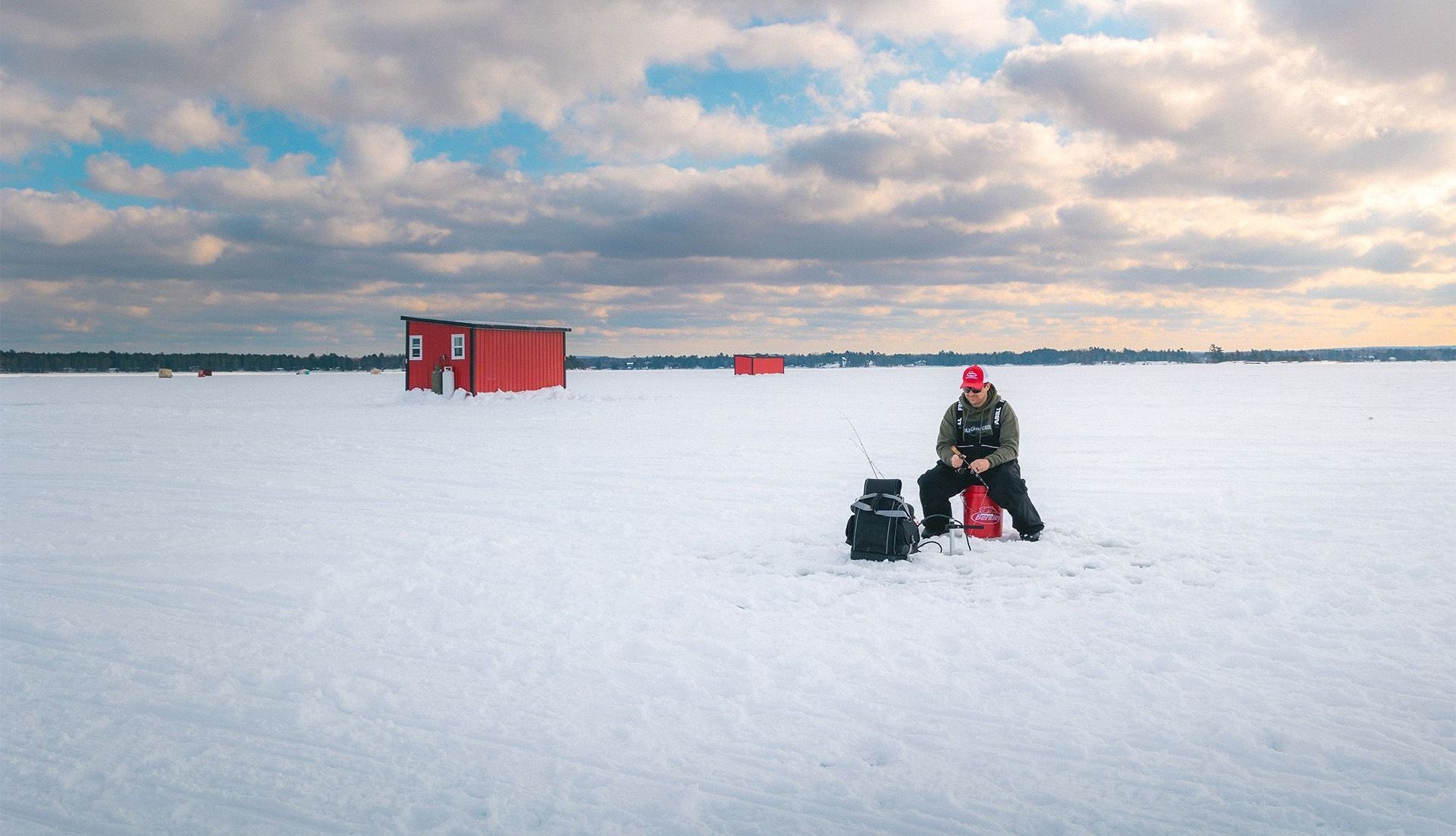 Une personne est agenouillée à côté d'un trou de glace. Au loin, on voit une cabane de pêche. Le ciel est immense, avec quelques percées bleues entre les nuages cotonneux.