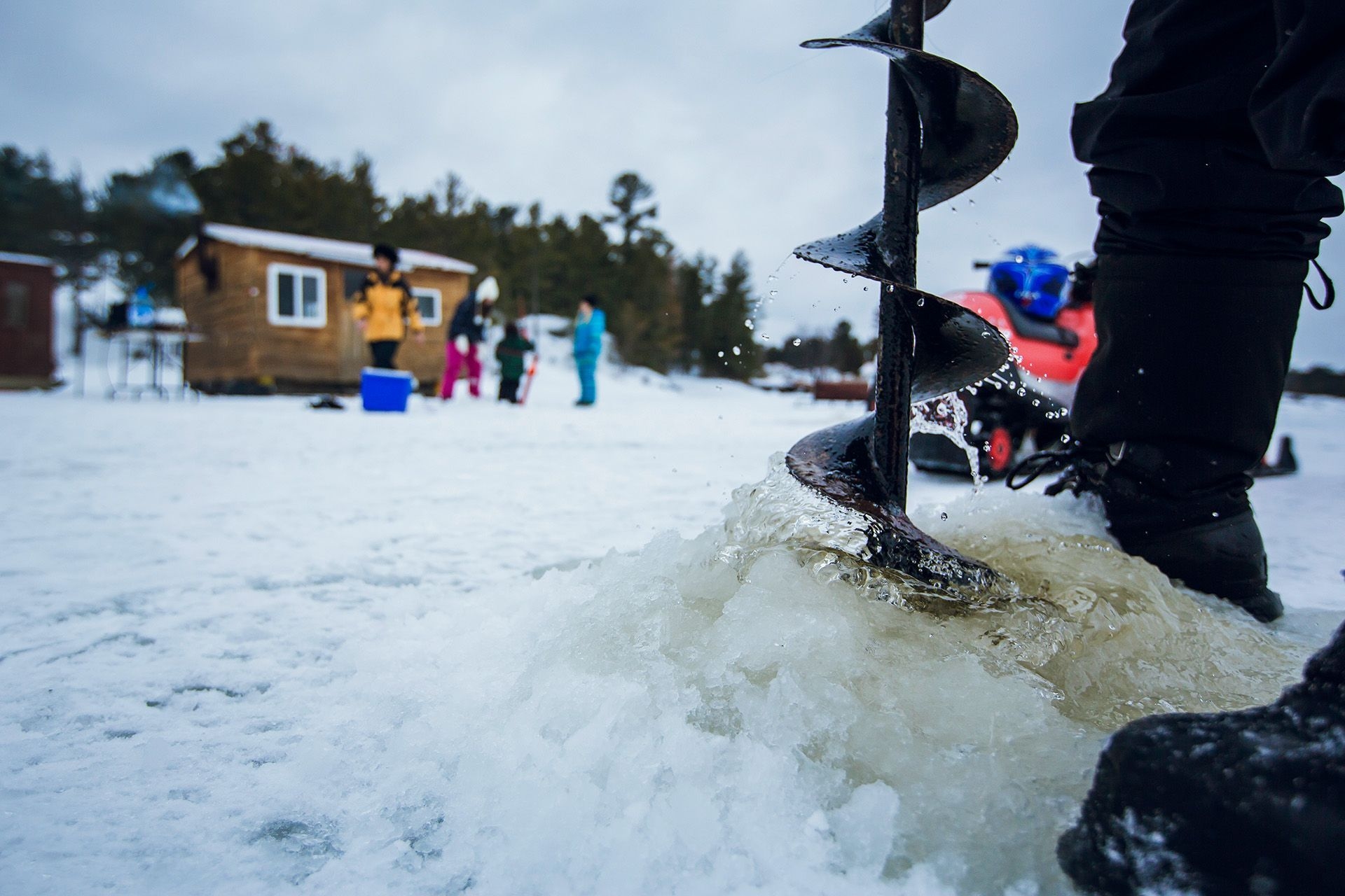 À l'aide d'une tarière à glace, une personne prépare un trou. Au loin, on aperçoit une cabane de pêche et un groupe. 