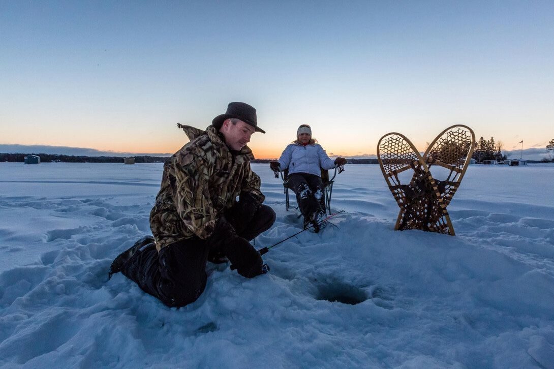 Une personne est assise sur une chaise de camping et observe un pêcheur, agenouillé près d'un trou de pêche. Des raquettes sont plantées dans la neige. 
