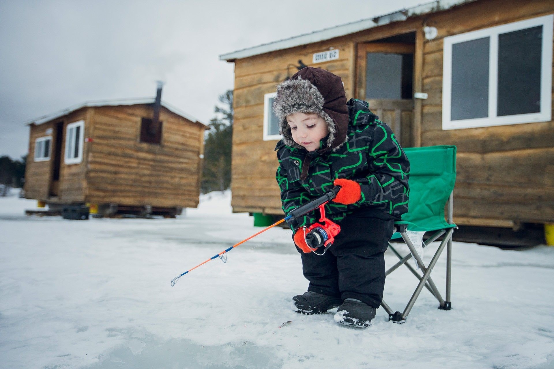 Un enfant d'âge préscolaire, qui porte un habit de neige et un chapeau, s'est levé les fesses de sa chaise de camping. Il est penché vers son trou de pêche. Il tient une canne a pêche. Il est installé devant des cabanes de pêche.