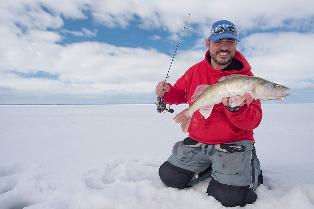 Un homme agenouillé tout près d'un trou de pêche blanche tient un gros poisson. Quel sourire!