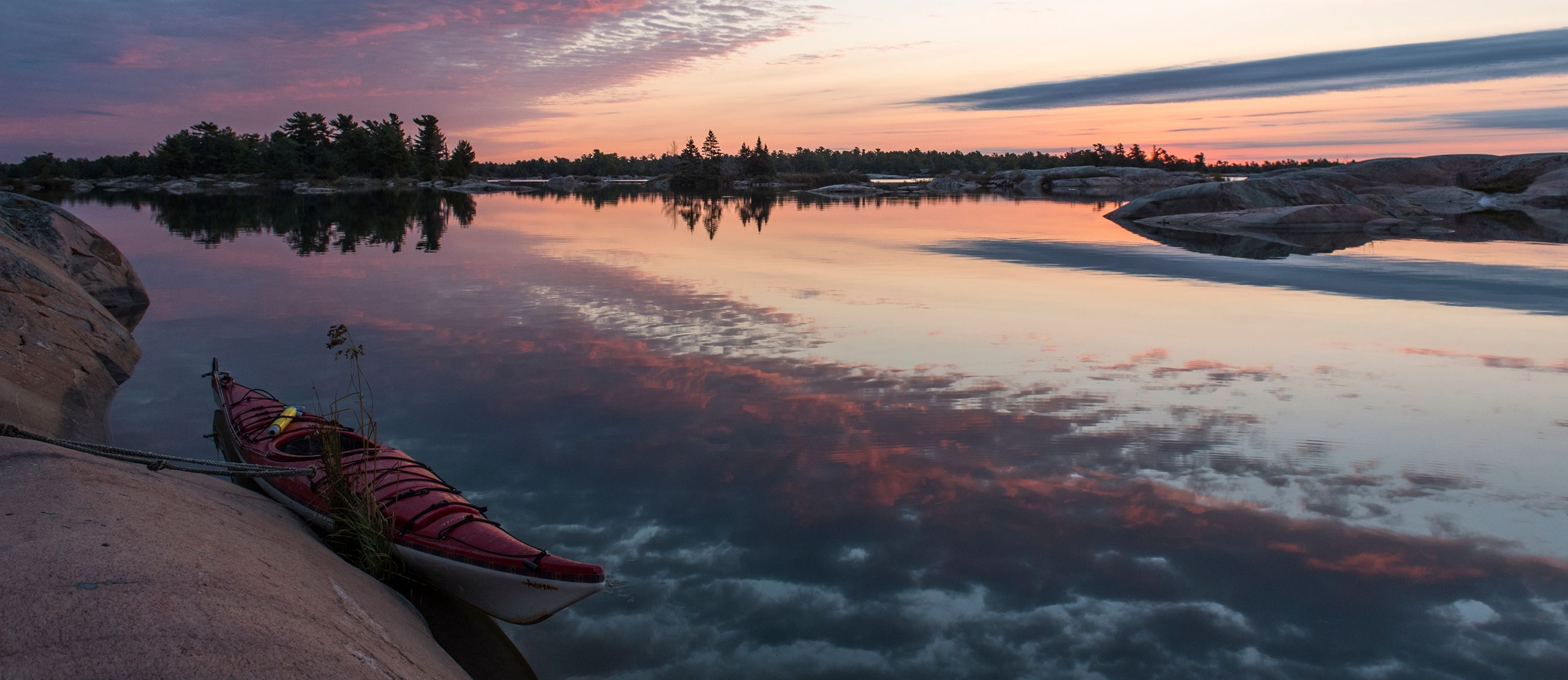 A kayak rests on a rocky shoreline of a perfectly still lake reflecting the pink and purple sunset