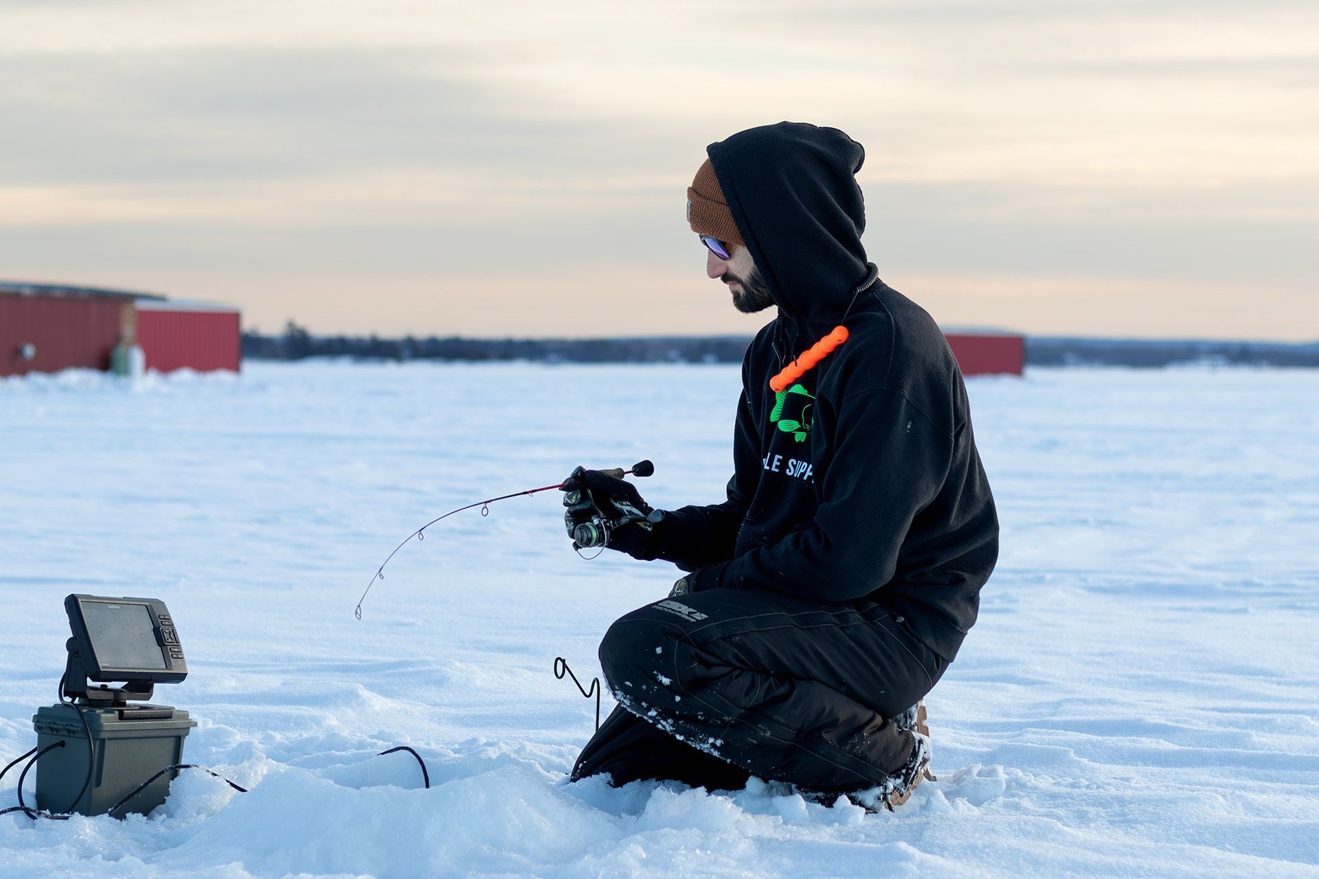 Un homme est agenouillé sur la neige, devant un trou de pêche. Il est emmitouflé. Le soleil semble être sur le point de se coucher. 
