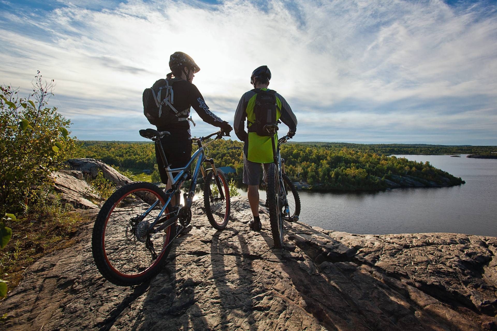 Deux adeptes du vélo de montagne portant un casque se tiennent sur un rocher et admirent un lac, à Sudbury