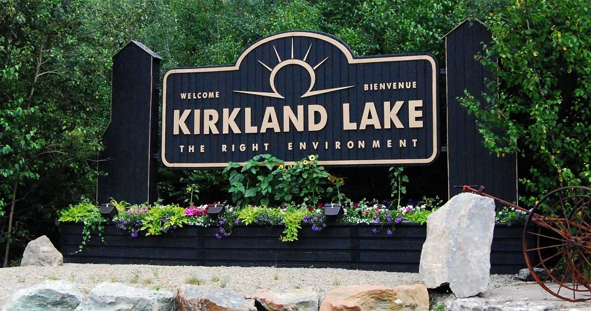 Le panneau de bienvenue de Kirkland Lake est foncé, avec le lettrage en jaune. Des bacs de fleurs et de verdure enjolivent l'endroit. 