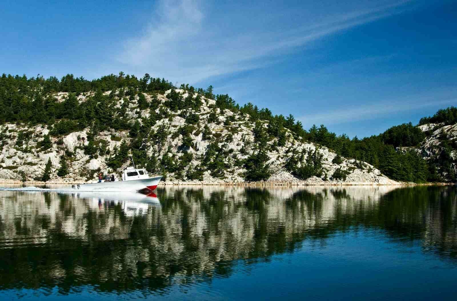 À l'arrière-plan, on voit une colline de granit, avec quelques arbres. La photo est prise à partir de l'eau. On voit un bateau à moteur circuler. 