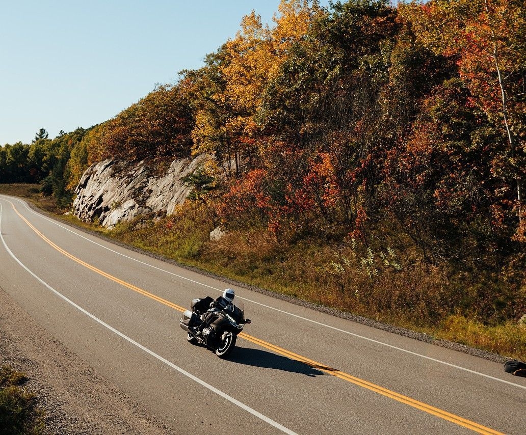 Un motocycliste, penché sur le moteur, s'apprête à négocier un virage. La route à double voie longe des rochers et des bosquets orange, jaune et rouge.