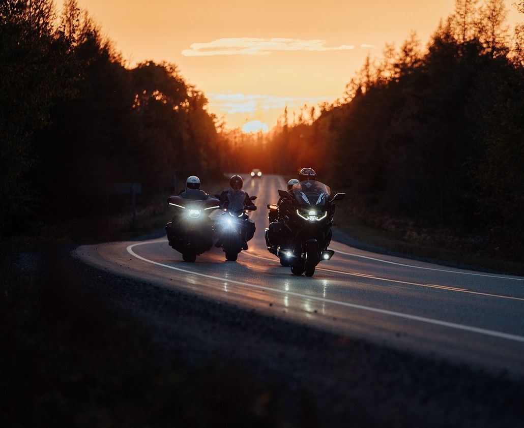 Trois motos circulent sur une route sinueuse. Elles font dos au soleil couchant.
