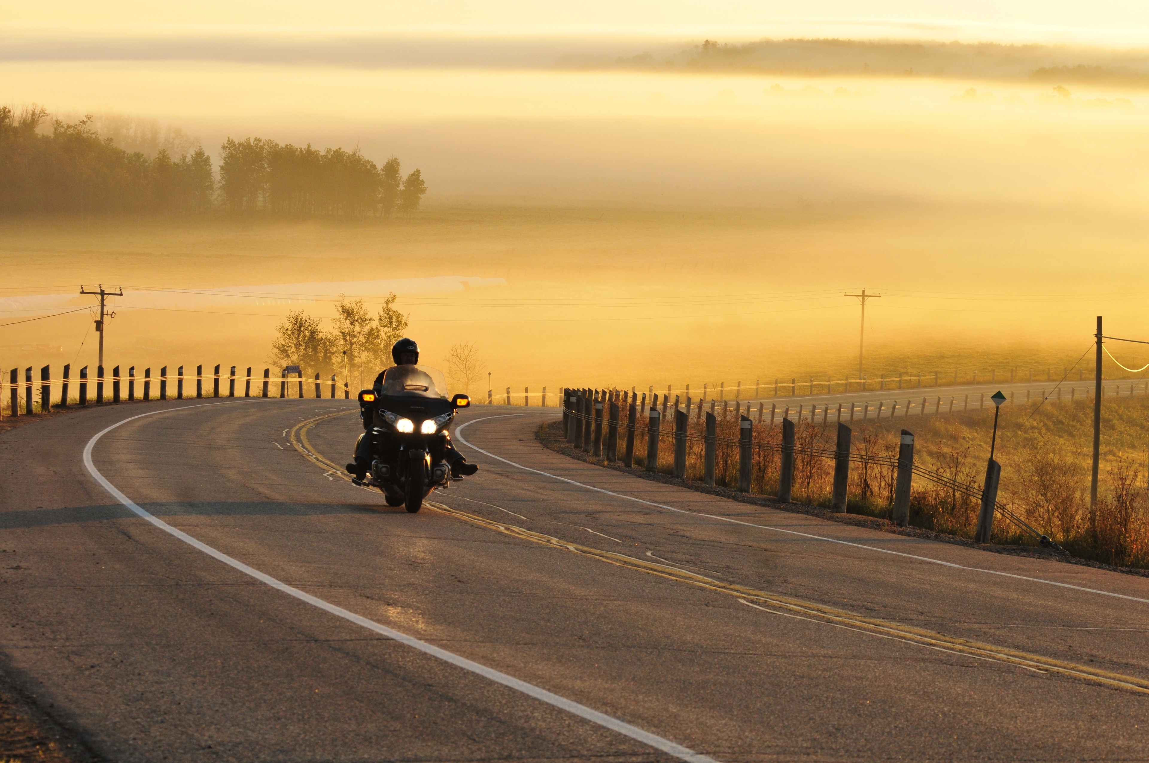 A biker drives on a curved road with a lake in the background reflecting yellow evening sunlight