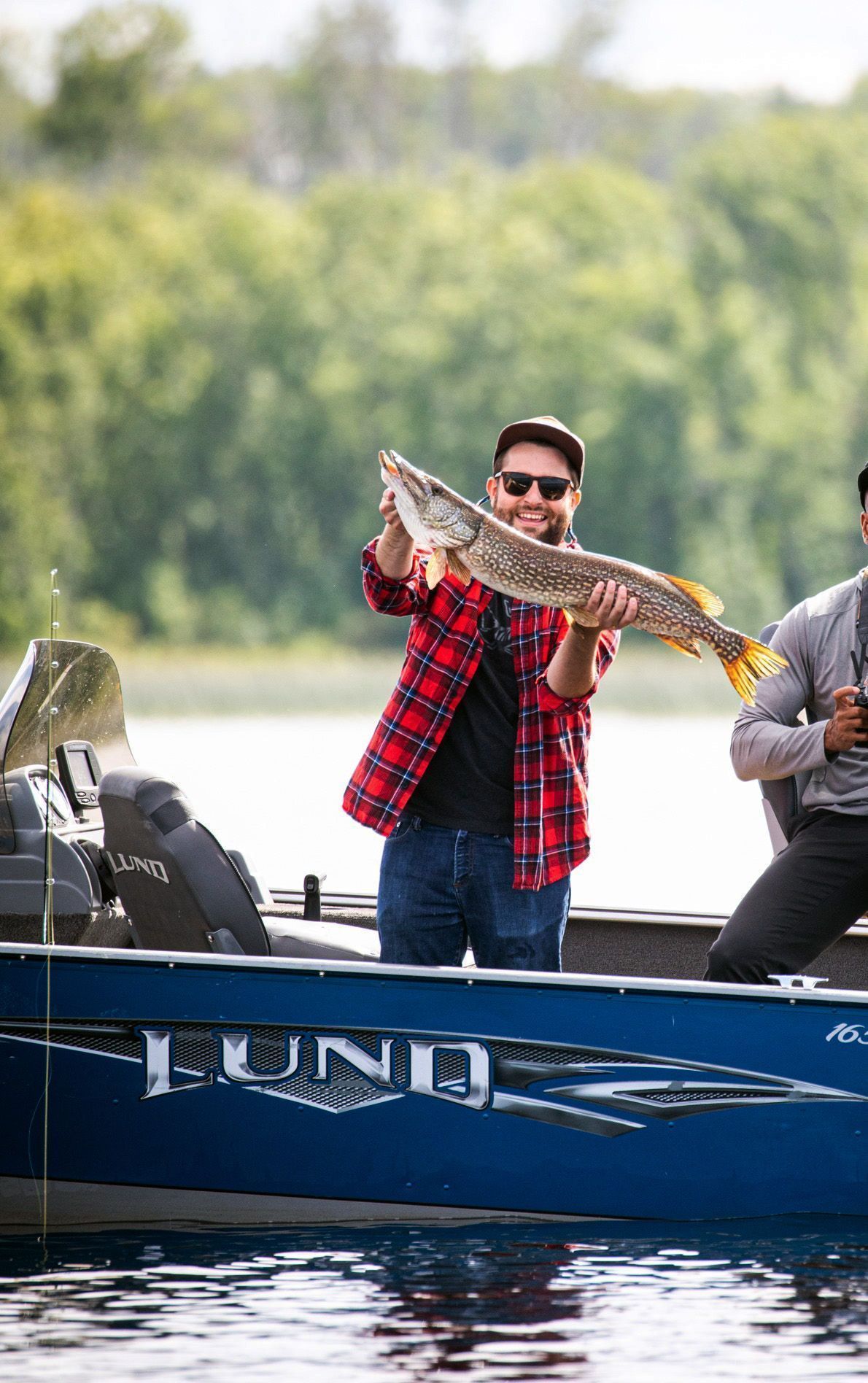 A young man in a red flannel shirt holds up a long pike he just caught.